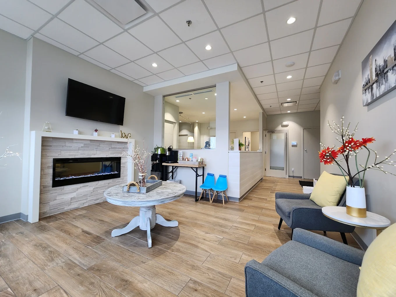 Modern waiting room with gray chairs, wooden floor, electric fireplace, TV, and decorative flowers.