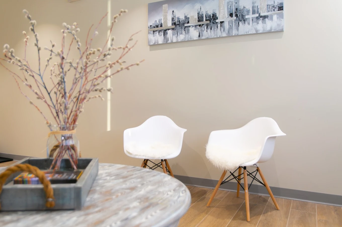 Two white modern chairs with wooden legs and fur cushions against a beige wall under a cityscape painting.