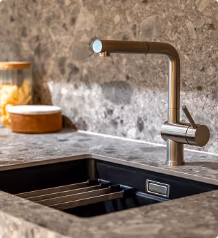Modern stainless steel kitchen faucet over a black sink set in a gray stone countertop and backsplash.