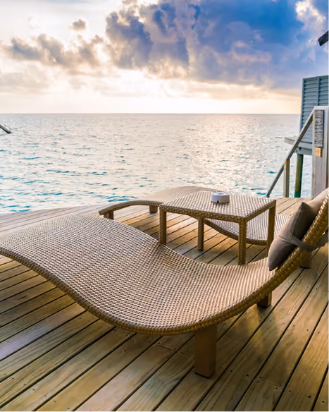 Wicker lounge chair and table on wooden deck overlooking calm ocean under cloudy sky.