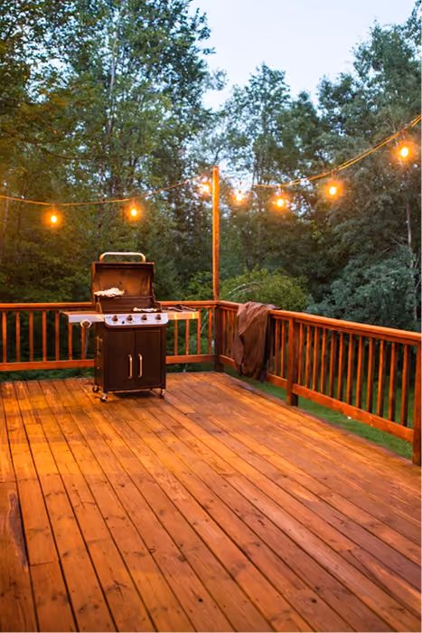 Wooden deck at dusk with string lights and a gas grill against a wooded background.