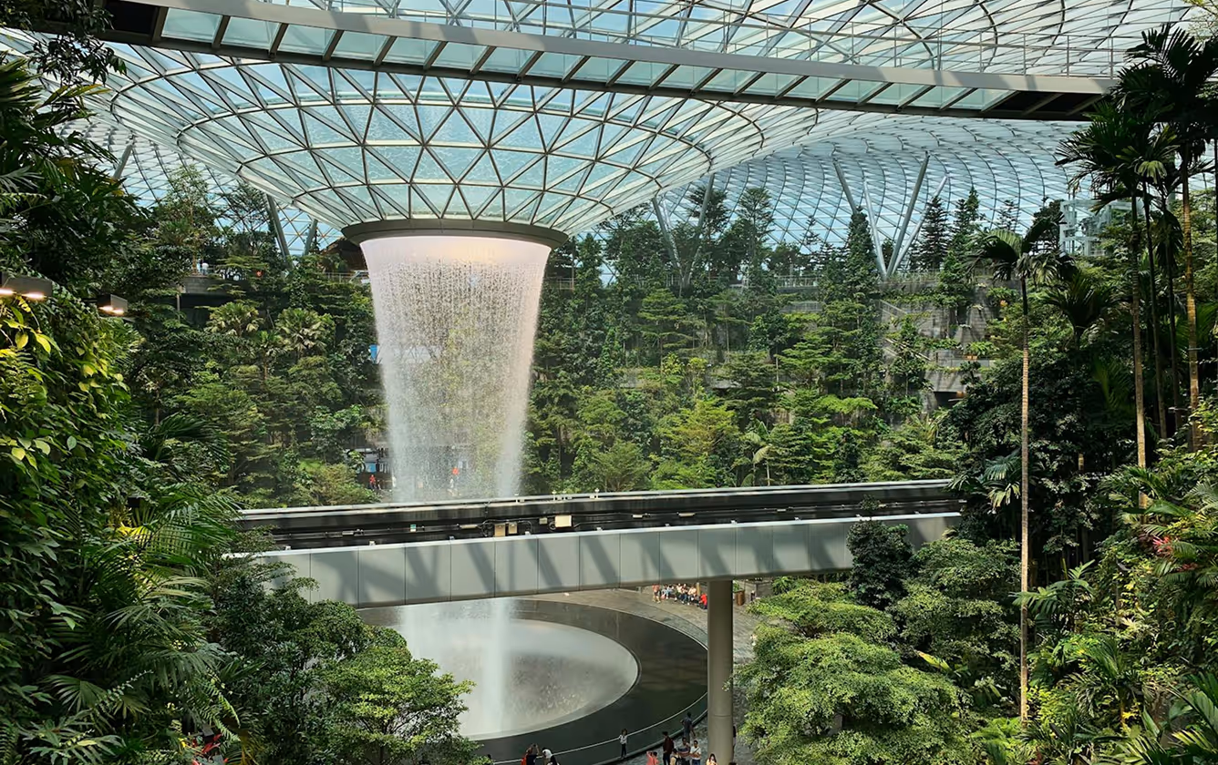 Indoor waterfall surrounded by lush greenery under a large glass dome ceiling with a walkway crossing above.