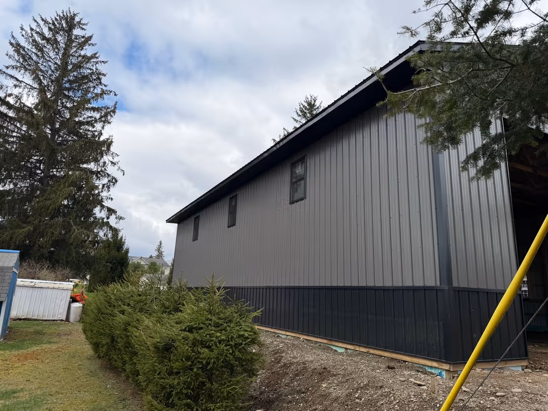 Side view of a gray metal building with three windows, surrounded by evergreen trees and bushes under a cloudy sky.
