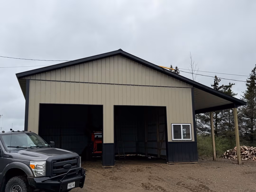 Metal barn with dual large open doors and a side window next to a dark gray pickup truck, with a pile of firewood and trees in the background under a cloudy sky.