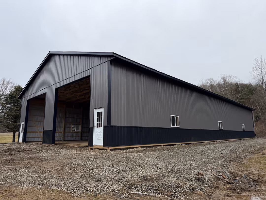 Large metal barn with open front bays, gray and black siding, white doors, and gravel ground around it on a cloudy day.