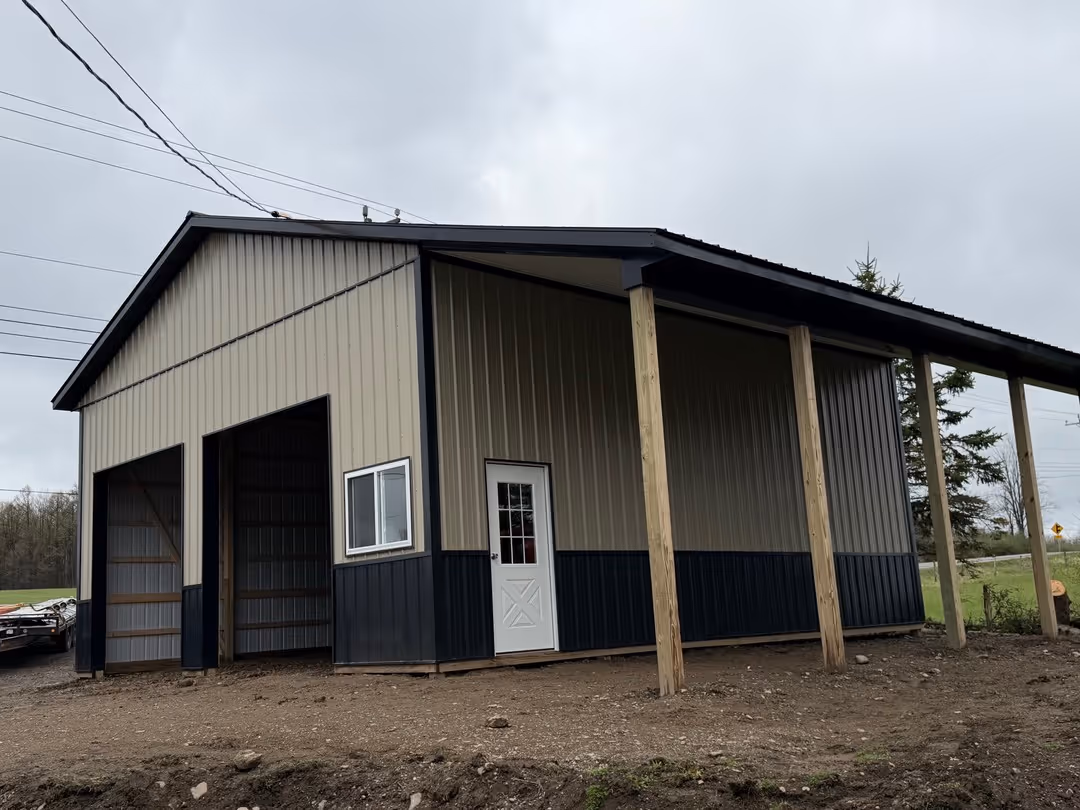 Metal barn with two large open bays, a small window, a white door, and a covered side supported by wooden posts.