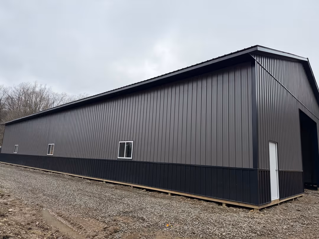 Large dark gray metal industrial building with a white door and three small windows on a gravel surface under a cloudy sky.