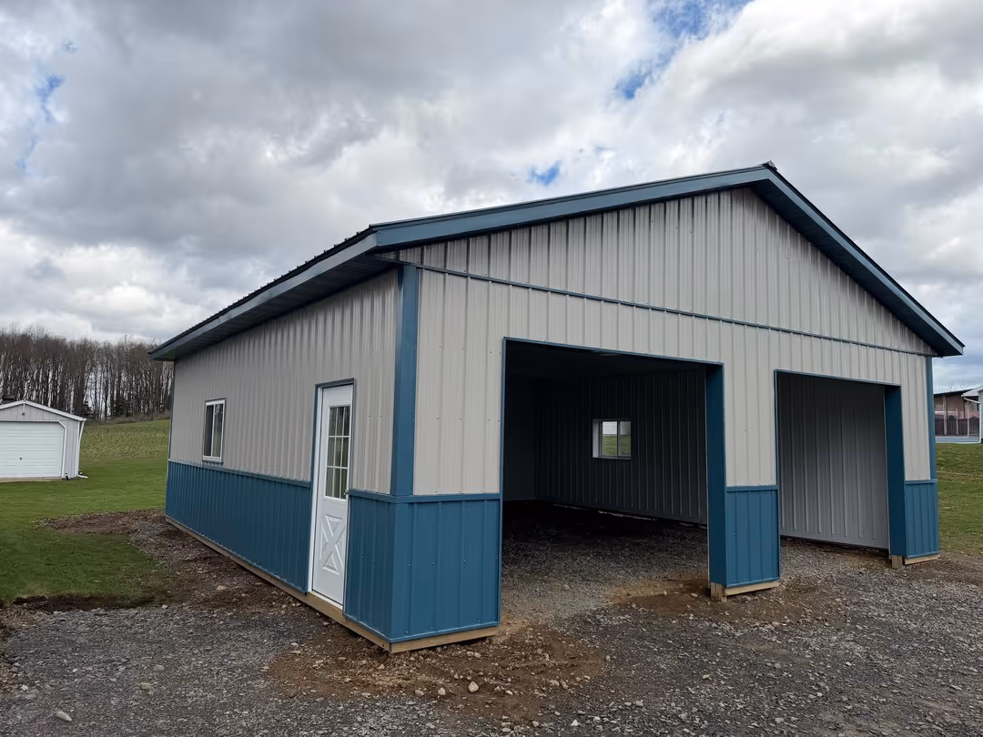 Metal carport with blue and gray panels, featuring a white side door and two open garage bays on a gravel driveway.