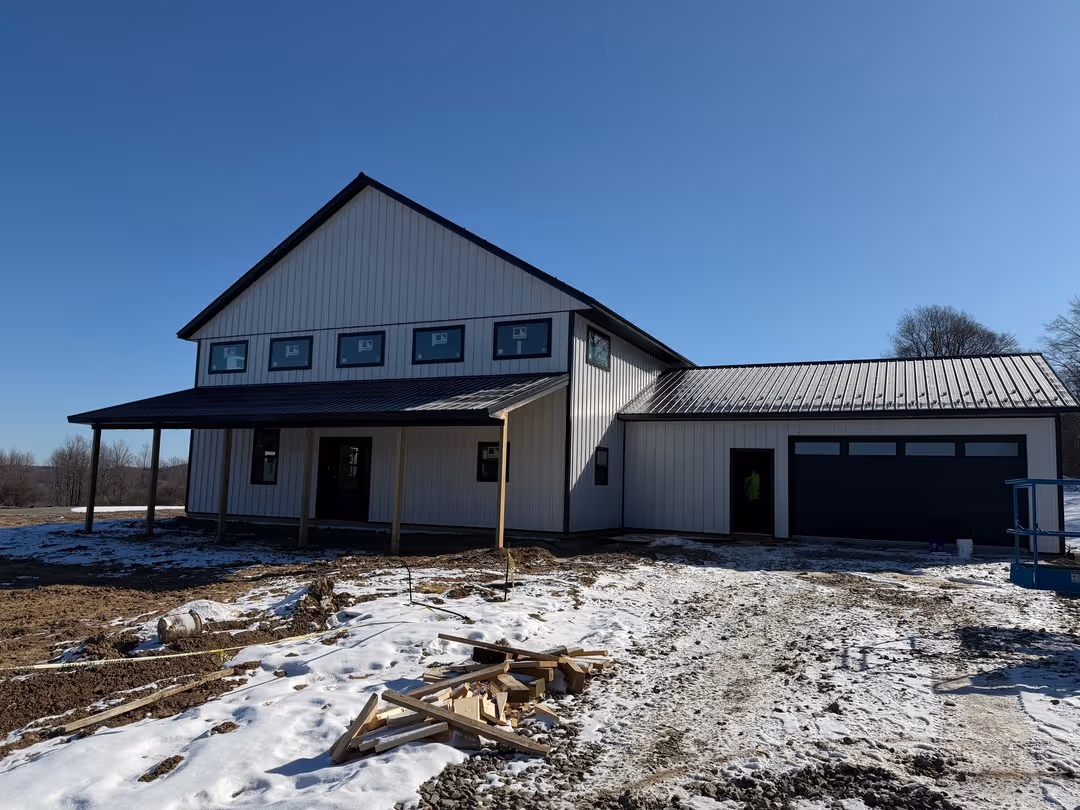 Modern two-story house under construction with white siding, black roof, and snow-covered ground in front.