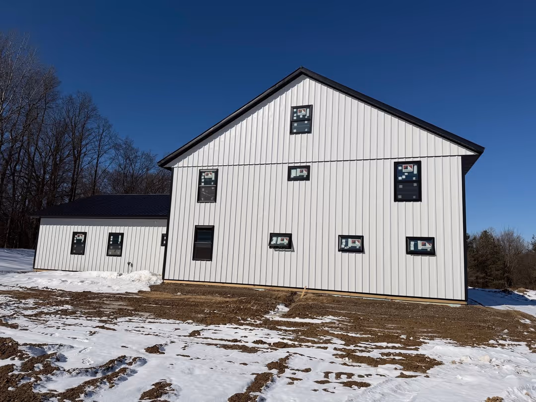 White modern barn-style building with multiple small black-framed windows, situated on a snowy and muddy ground with leafless trees in the background under a clear blue sky.