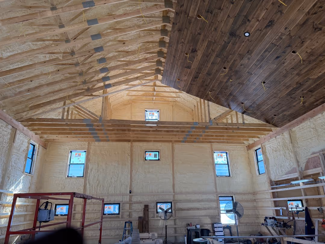 Interior view of a large room under construction with exposed wooden roof trusses, unfinished insulation on walls, and partially installed dark wood ceiling paneling.