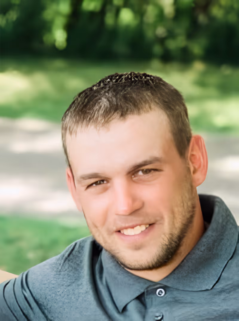 Smiling man with short brown hair and light stubble wearing a gray collared shirt outdoors with greenery in the background.