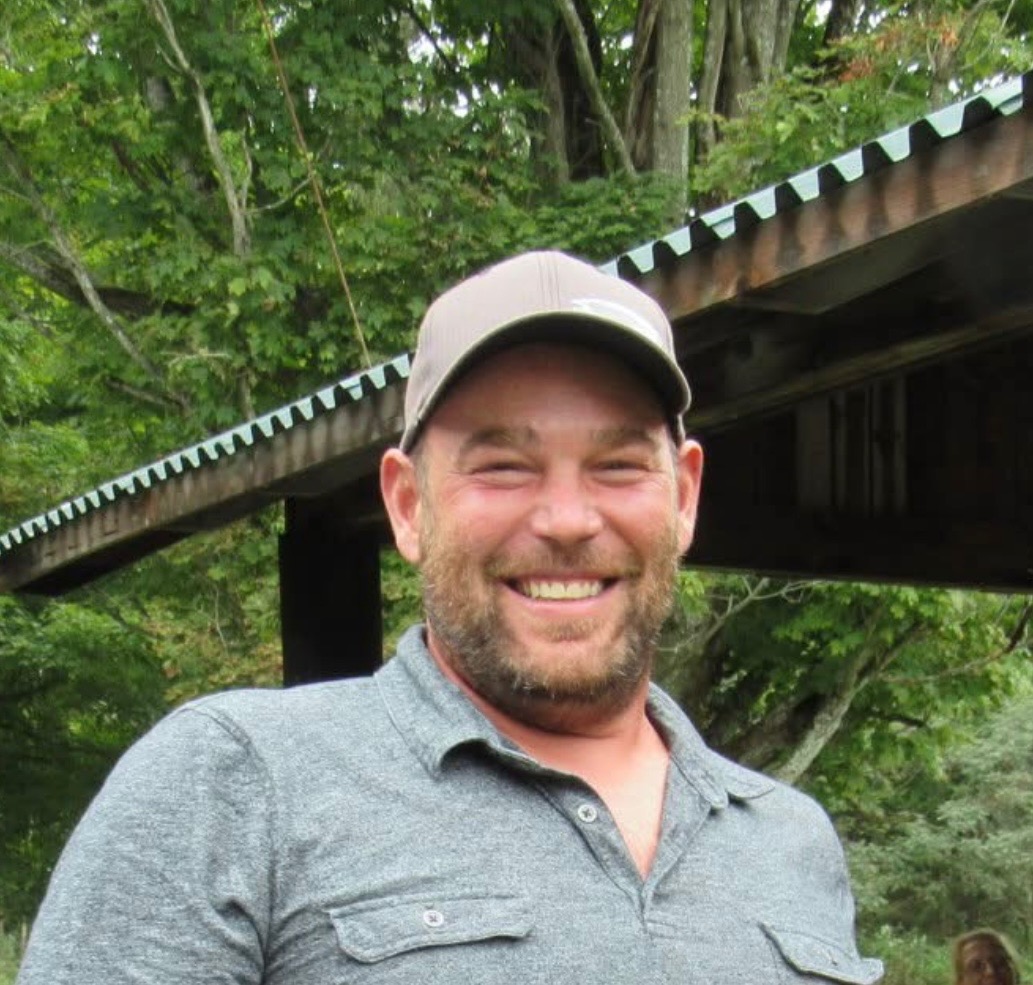 Smiling man with beard wearing gray cap and shirt standing outdoors near wooden structure and trees.
