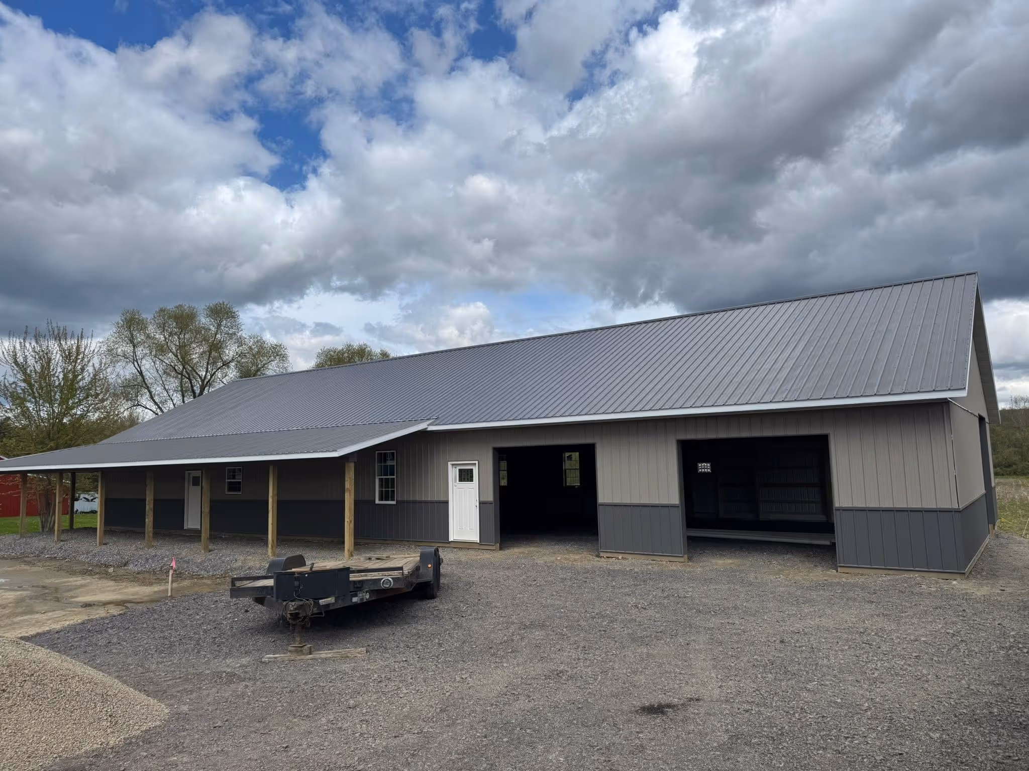 Large gray metal barn with two open bays, white door, and covered porch on gravel ground under cloudy sky.