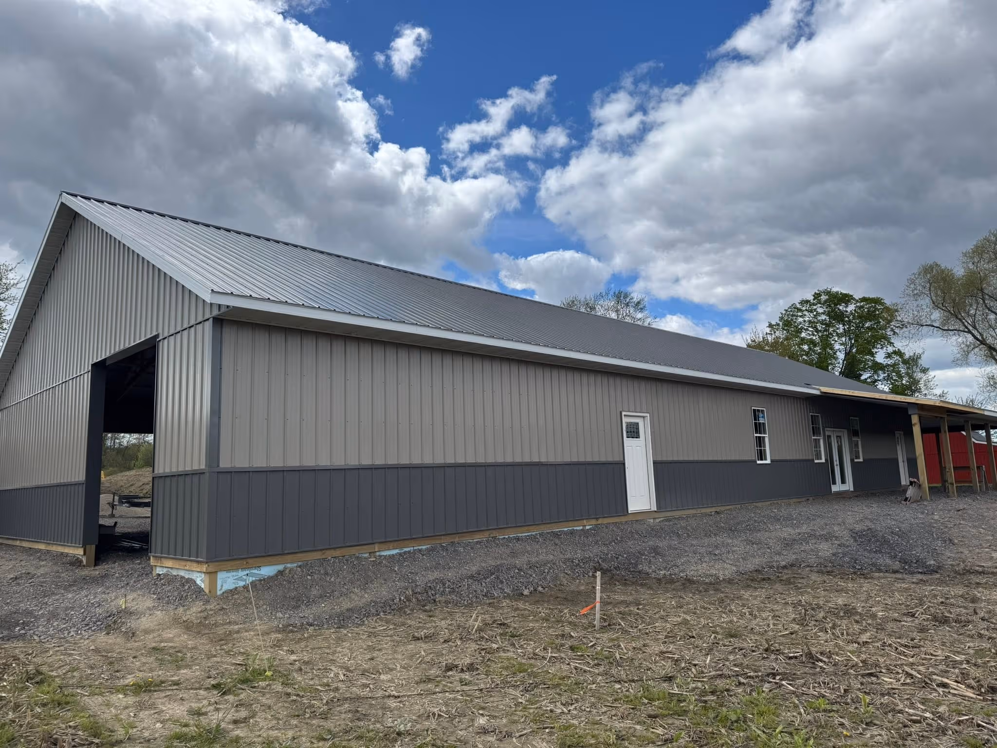 Large gray metal barn with white doors under a partly cloudy sky on a gravel and dirt lot.