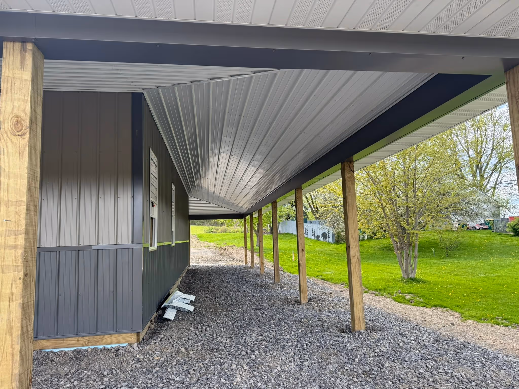 Covered carport with wooden posts beside a metal-sided building on a gravel surface with grass and trees beyond.