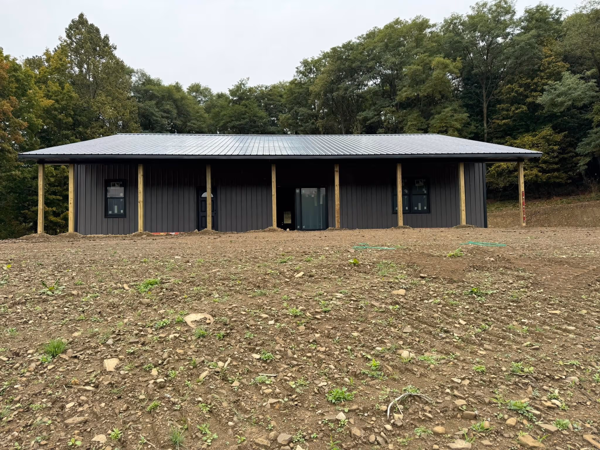 New gray metal building with a metal roof on cleared rocky land, surrounded by dense green trees.