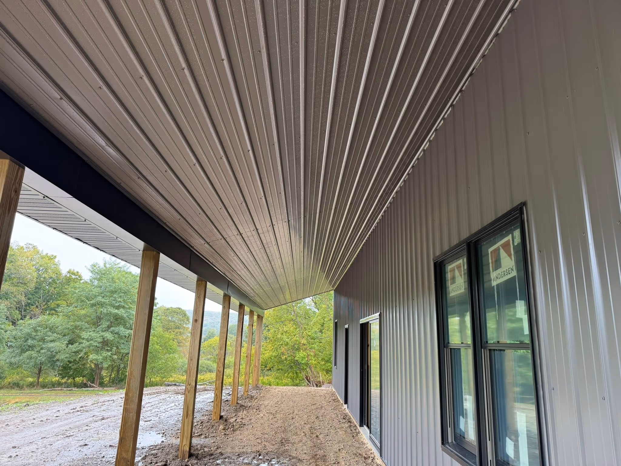 Side view of a building with a metal awning supported by wooden posts and trees in the background.