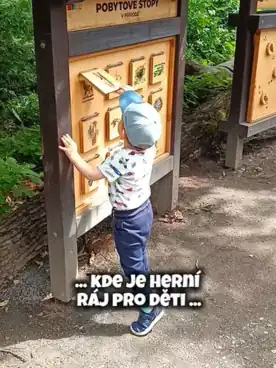 Dítě u naučné tabule v lese s nápisem 'Kde je herní ráj pro děti'.