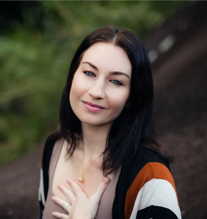 Woman with dark hair and a nose ring smiling gently outdoors, wearing a striped cardigan and a necklace, with hand placed on her chest.