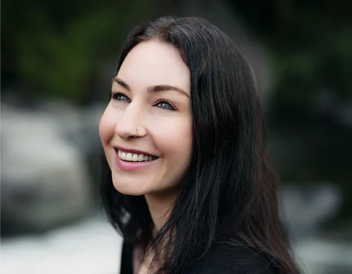 Smiling woman with long dark hair, blue eyes, and a nose ring looking upward against a blurred outdoor background.