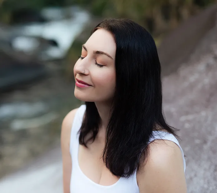 Woman with long dark hair wearing a white tank top, eyes closed and smiling peacefully outdoors.