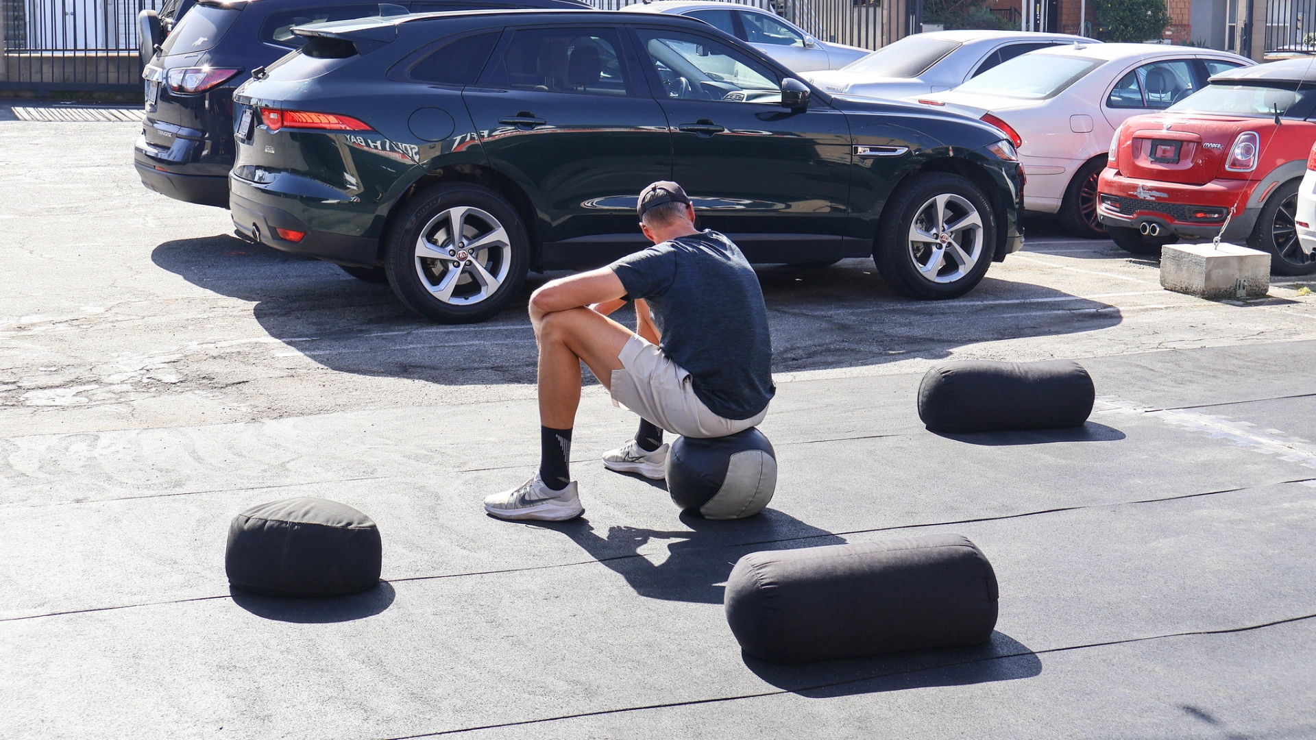 A man rests on a medicine ball in an outdoor workout area surrounded by sandbags and parked cars. The sunny training space outside the gym creates a casual recovery moment between exercises.