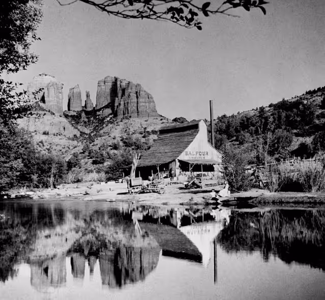 Black and white photo of Balfour Mill by a pond with mountain cliffs reflected in the water.
