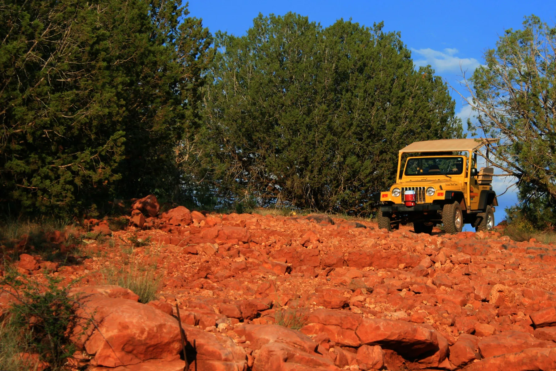 Yellow Jeep on a rocky red dirt trail surrounded by green trees under a blue sky.