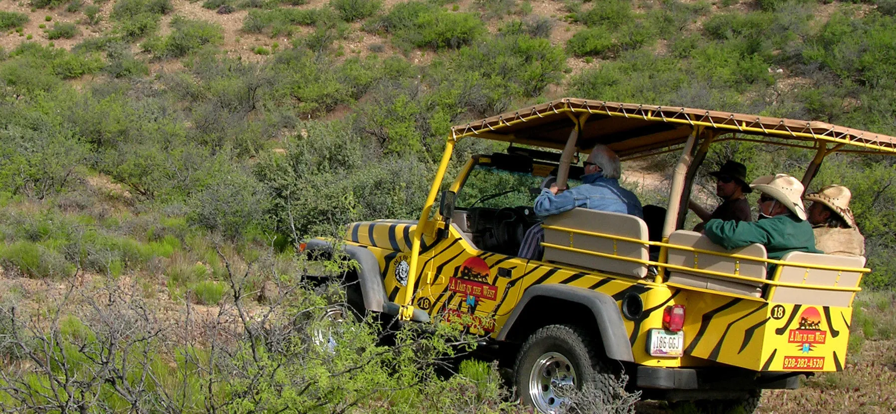 Yellow safari jeep with passengers wearing hats driving through a green desert landscape.