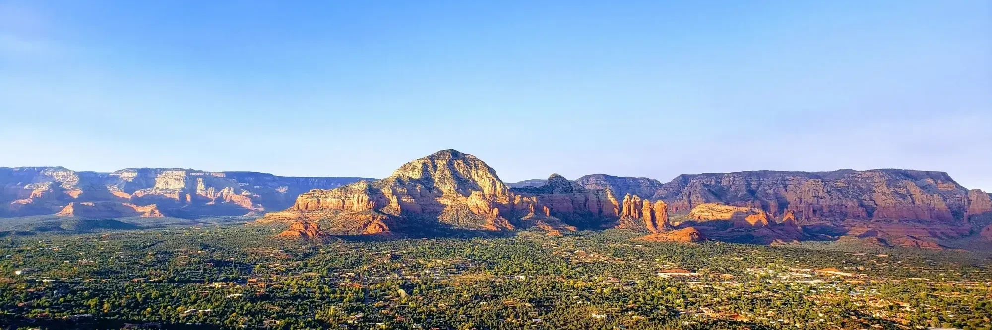 Panoramic view of red rock mountains and green forest under clear blue sky at sunset.
