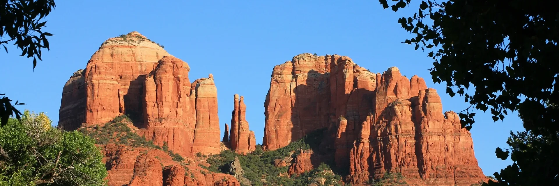 Red rock formations in a desert landscape under a clear blue sky framed by leafy tree branches.