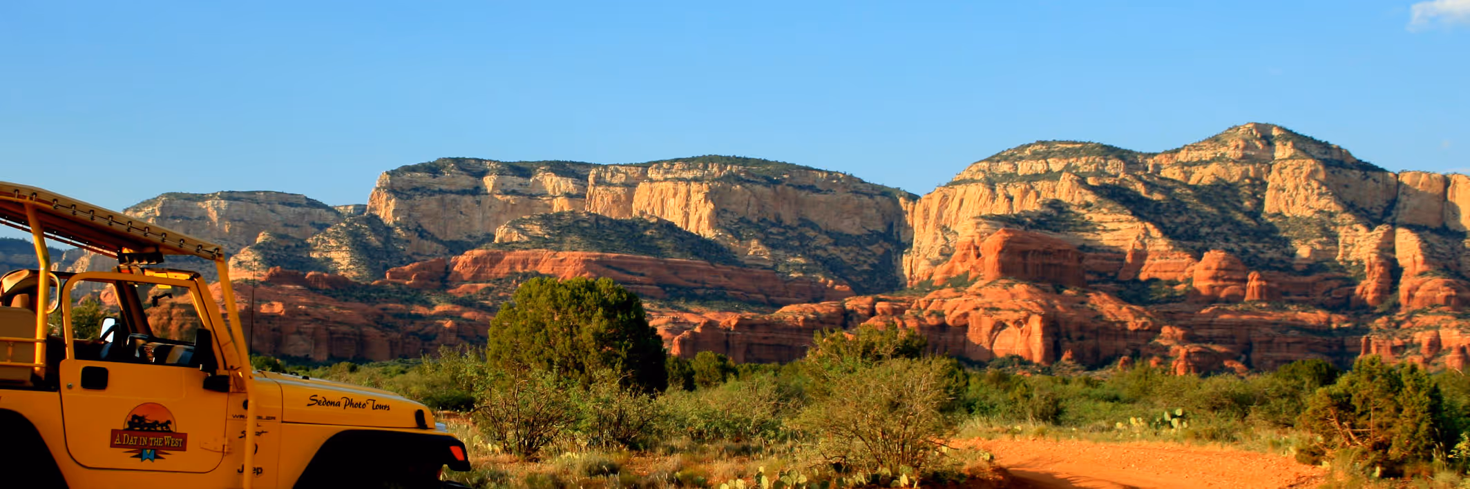 Yellow jeep labeled 'A Day in the West' parked near red rock formations and desert vegetation under a clear sky.