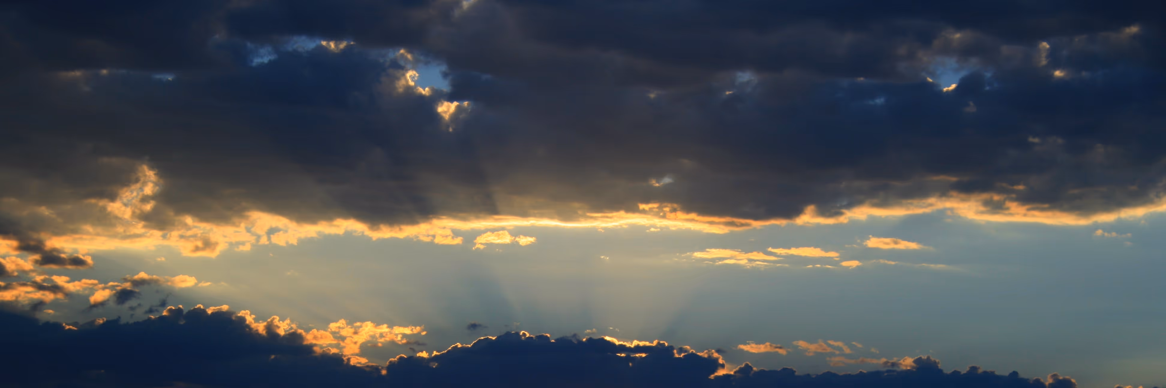 Sunset sky with dark clouds and golden light rays shining through gaps in clouds.