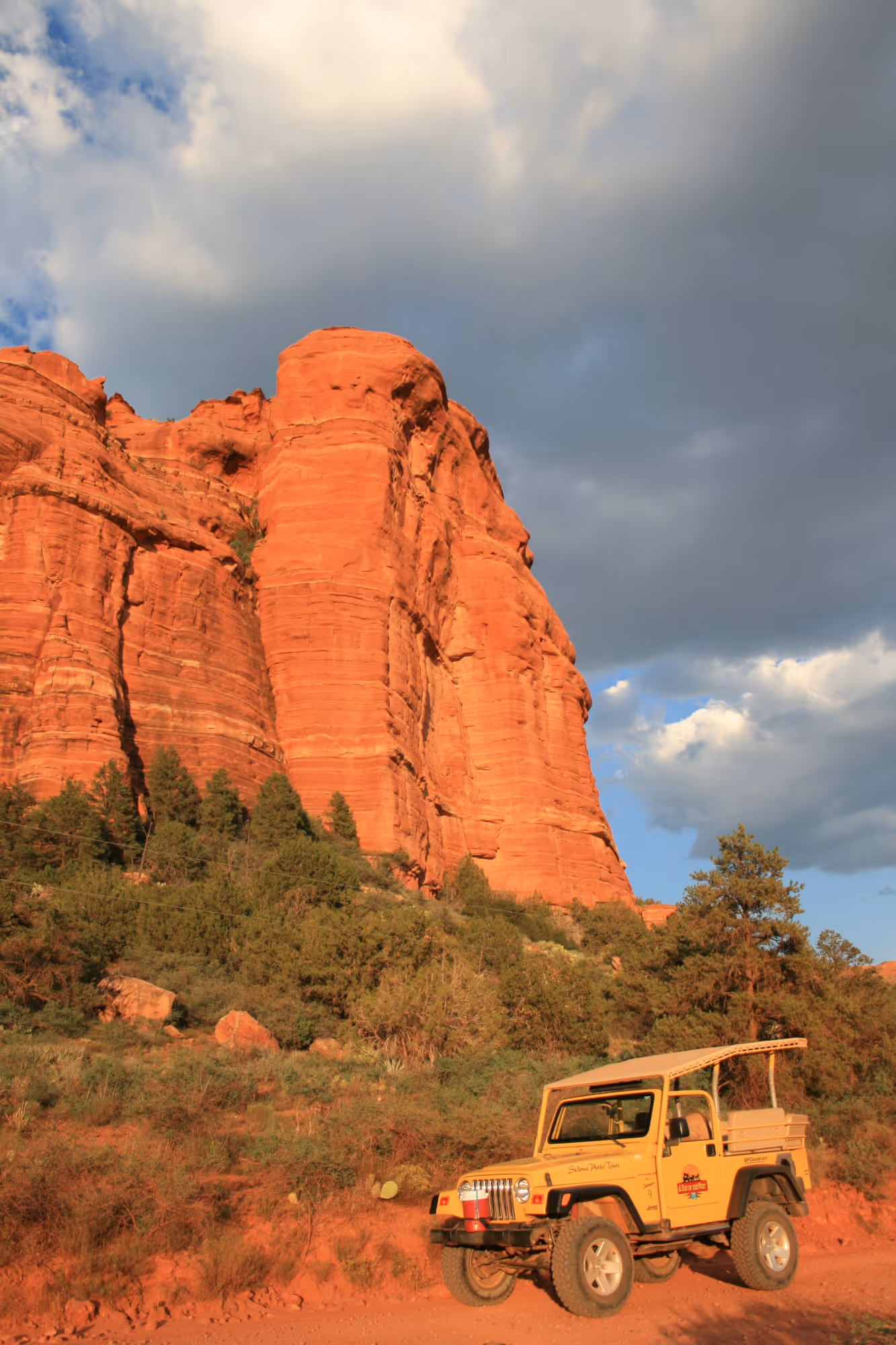 Yellow jeep parked on a red dirt road with towering red rock formations and green trees nearby.