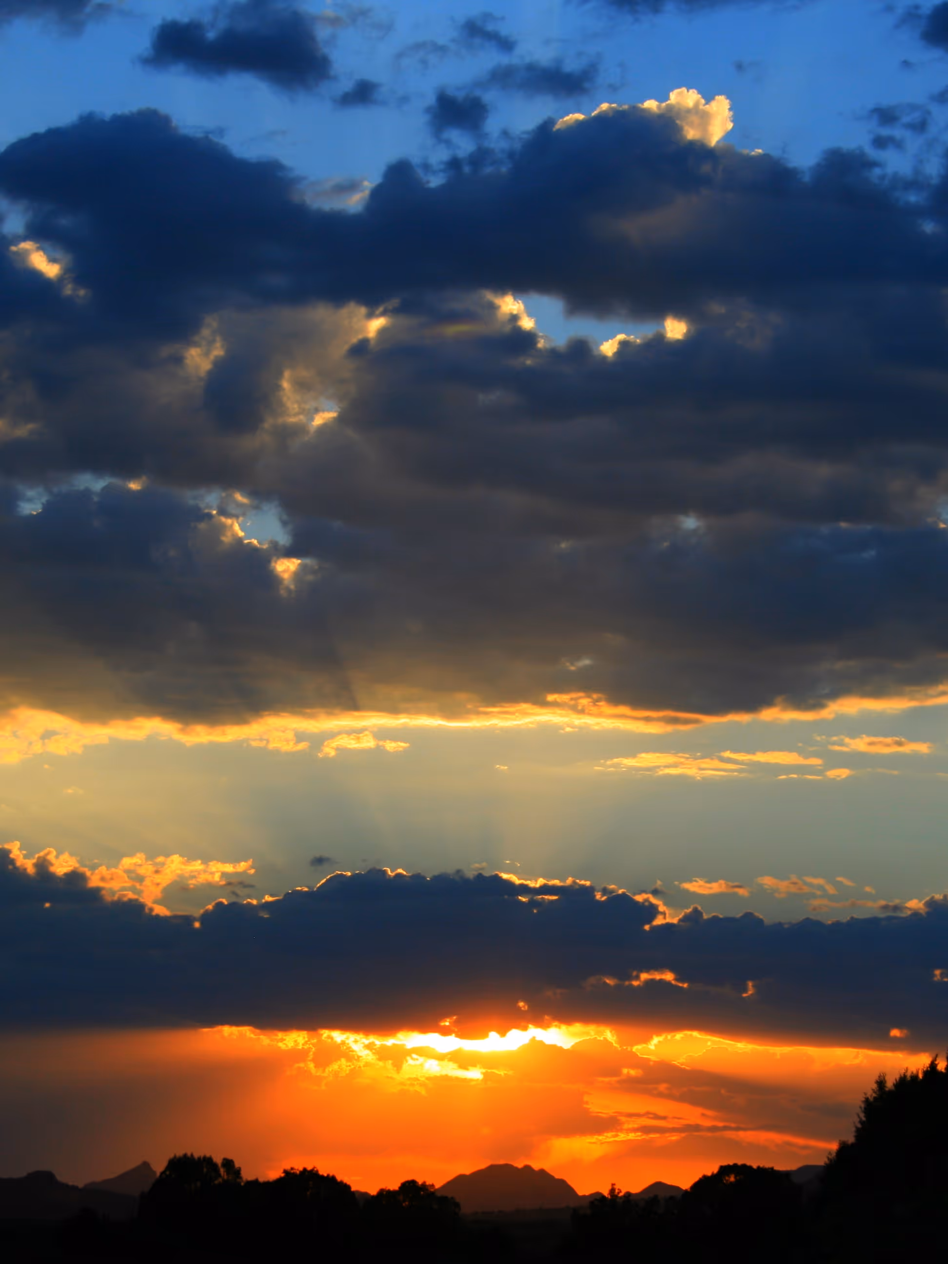 Sunset with bright orange light peeking through dark clouds over distant mountains and trees.