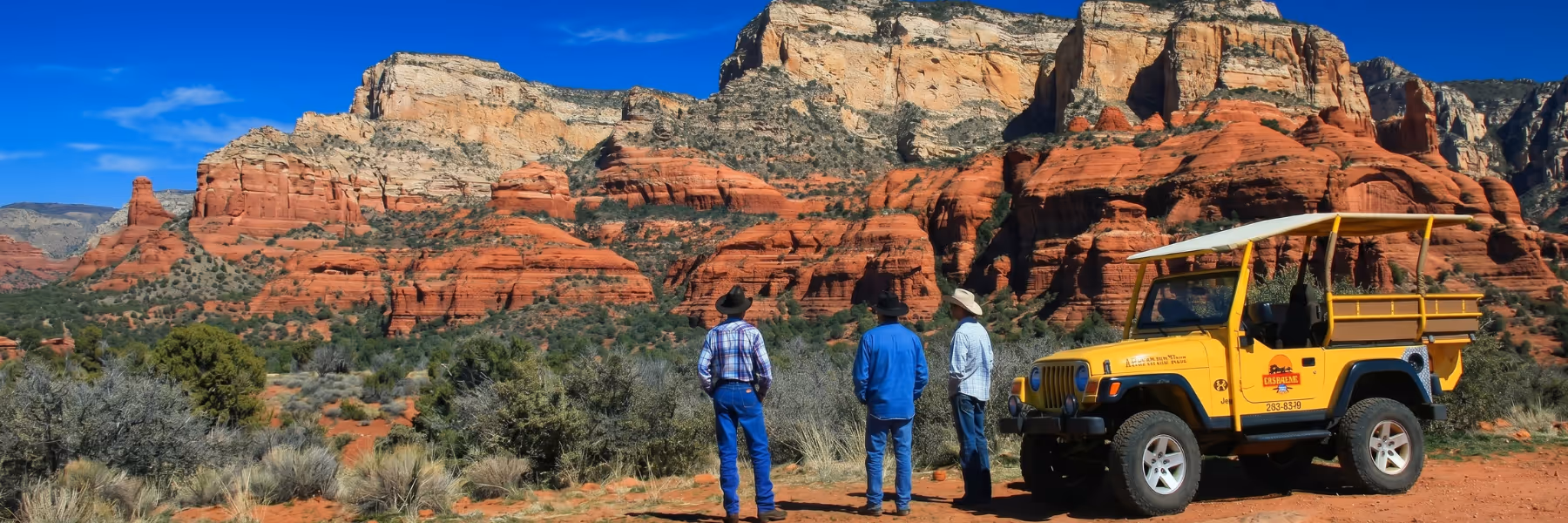 Three men in cowboy hats stand near a yellow jeep with red rock formations under blue sky.