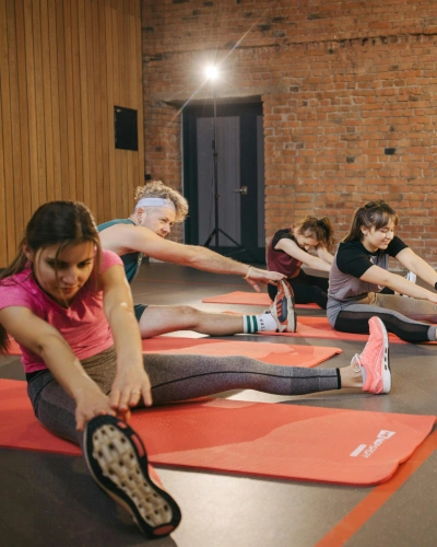 A group of five people stretches on red mats in a gym with brick walls. They reach forward, touching their toes, focused and relaxed.