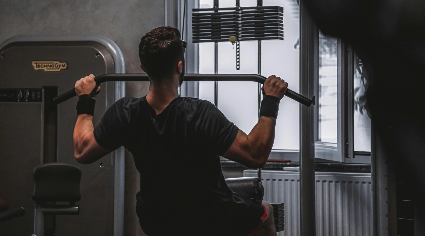 A person with short hair performs a seated lat pulldown in a gym. Wearing a black shirt, they grasp a bar, showing strength and focus. Equipment and a large window are visible.