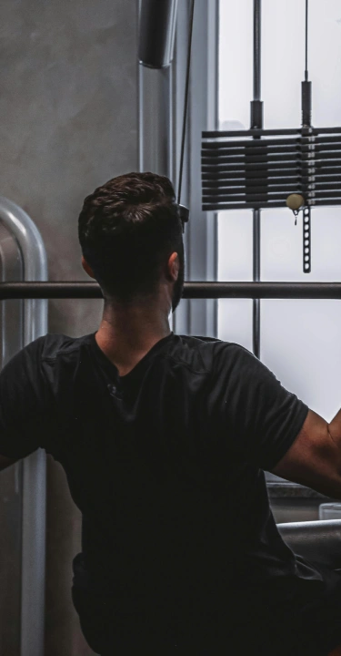 A person with short hair performs a seated lat pulldown in a gym. Wearing a black shirt, they grasp a bar, showing strength and focus. Equipment and a large window are visible.