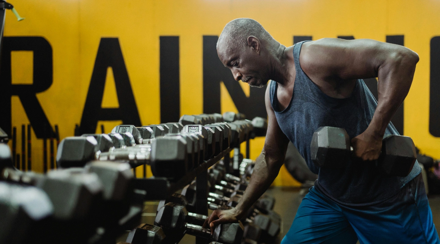 Man lifting dumbbells in a gym, wearing a tank top and blue shorts. Yellow wall with large letters behind him conveys focus and intensity.