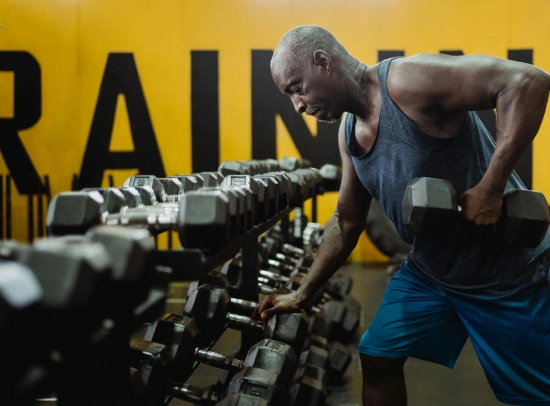Man lifting dumbbells in a gym, wearing a tank top and blue shorts. Yellow wall with large letters behind him conveys focus and intensity.