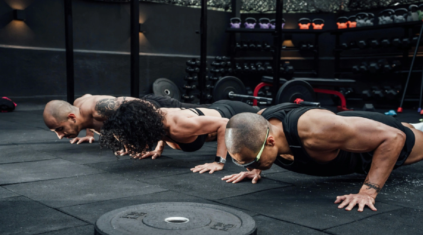Three individuals perform push-ups on a gym's black rubber floor, surrounded by weights and equipment, showcasing strength and focus.