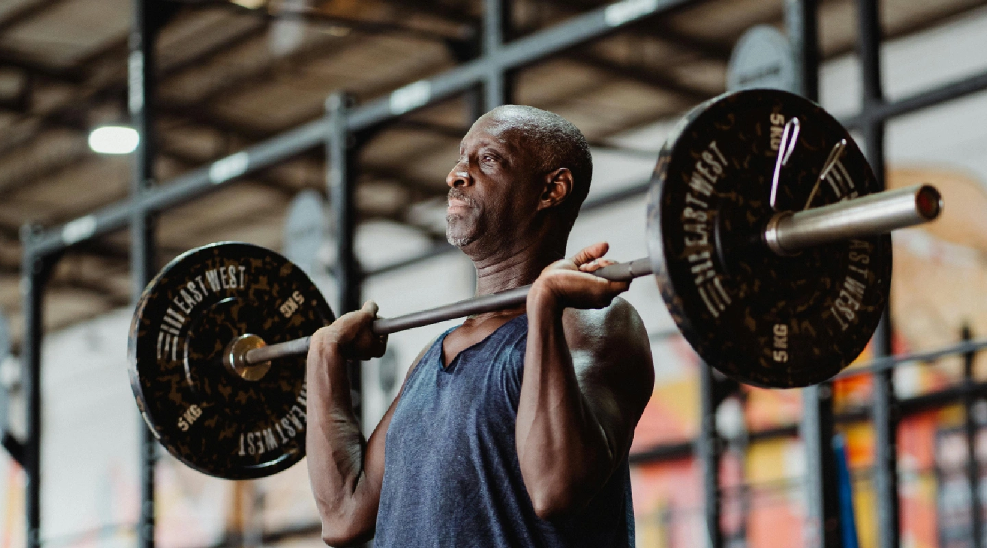 A man in a gym lifts a barbell with focus and determination. He wears a sleeveless shirt, surrounded by gym equipment. The scene conveys strength and endurance.