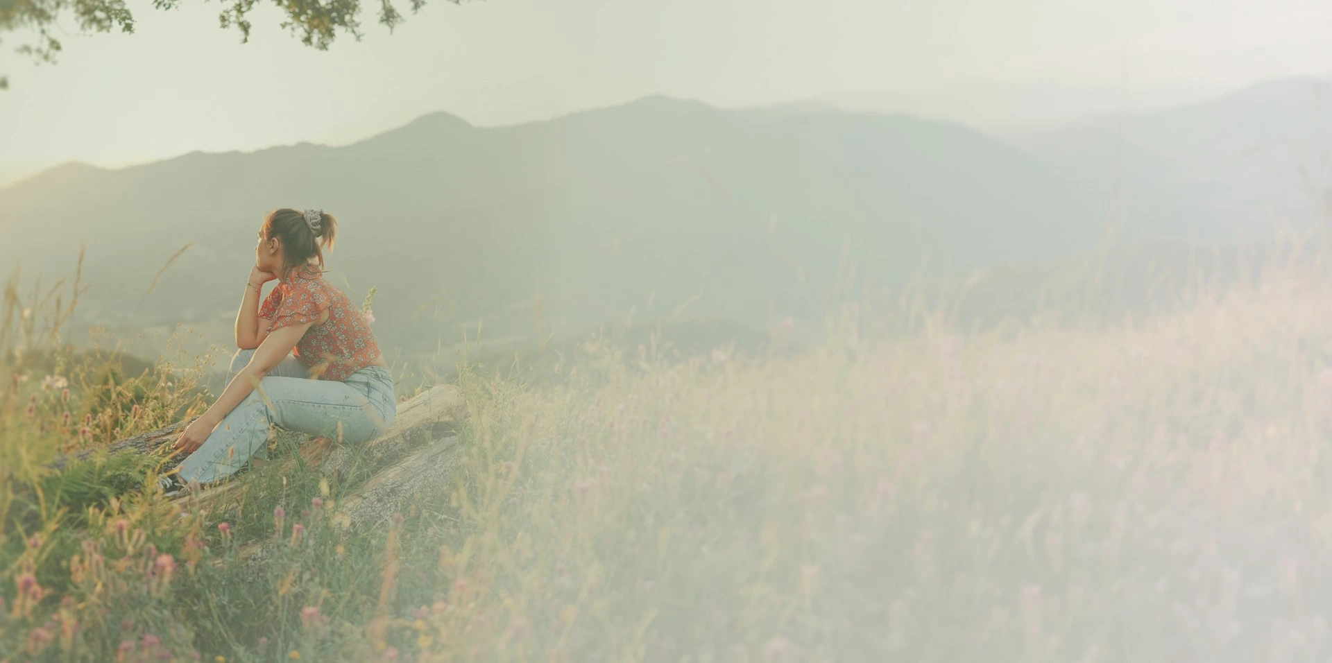 Woman in a red floral shirt and light jeans sitting on a log in a meadow with mountains in the background.