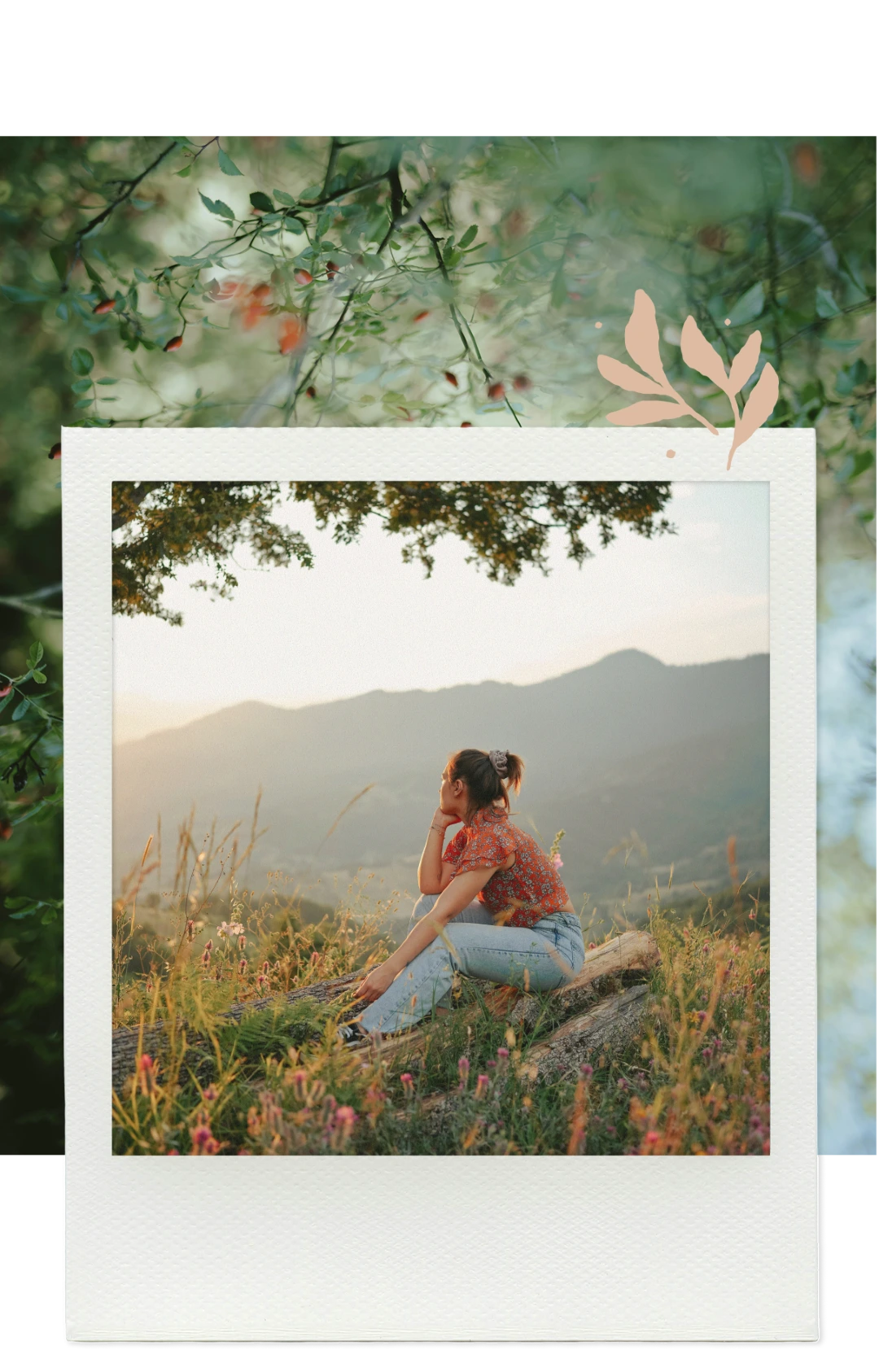 Woman in a red floral top and light jeans sitting on a log in a grassy field with mountains in the background during sunset.