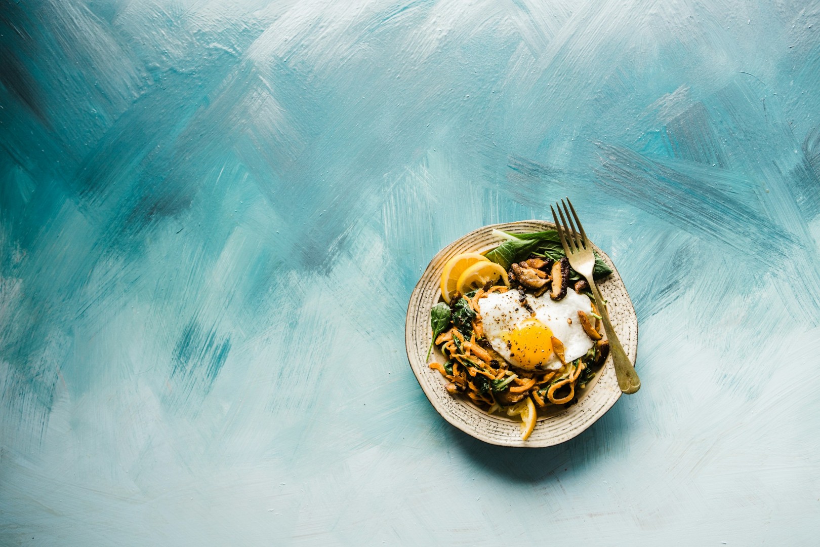 Plate with fried egg, sautéed mushrooms, lemon slices, and vegetable noodles on blue textured surface.