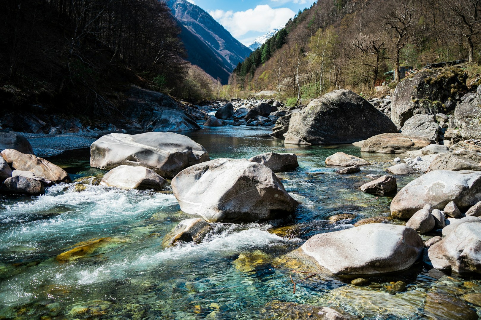 Clear river with large rocks flowing through a mountainous forested landscape under blue sky.