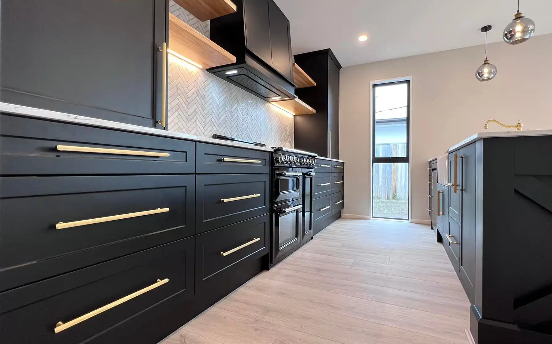 Modern kitchen with black cabinetry, brass handles, light wood shelves, and a window at the end.