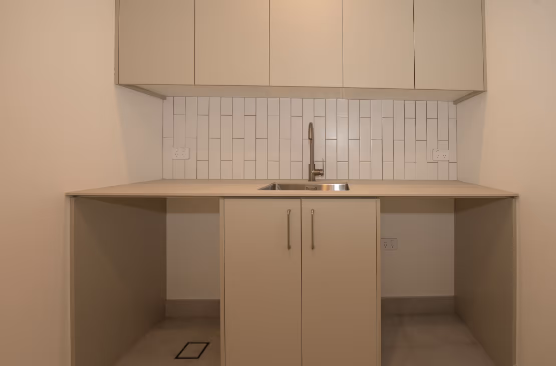 Minimalist beige laundry room with a sink, upper cabinets, white subway tile backsplash, and two open spaces below